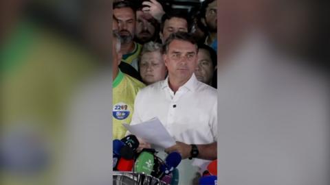 Senator Flavio Bolsonaro wearing white shirt and speaking in front of row of mics