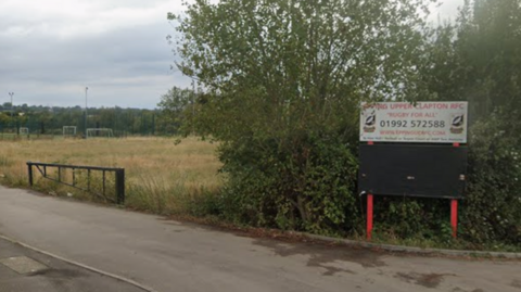 A narrow entrance road to a rugby club. A white sign, with red font, is at the entrance. It reads "EPPING UPPER CLAPTON RFC". There is a field with long grass to the left of the frame.