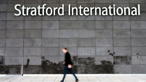 A man who is blurred walks past a grey wall with white lettering that reads Stratford International.