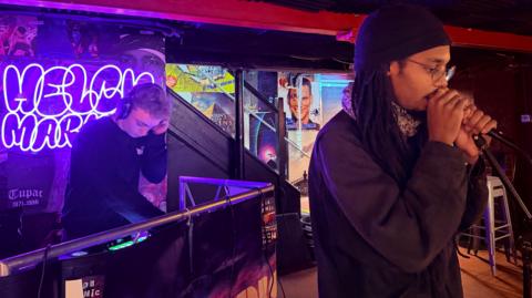 Yannick is mid-performing at a microphone, with Jack in the background DJing and mixing music. Yannick is wearing a dark beanie over his locs. Jack is wearing headphones. Behind Jack is a glowing, neon purple 'Helch Market' sign.