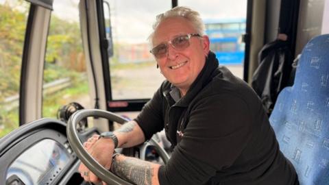 A bus driver leaning over the wheel in his cab whilst parked up. He is looking at the camera smiling. He has grey hair and is wearing slight tinted glasses with a silver frame. He also has a black fleece on with the sleeves rolled up showing off tattoos on his forearms.