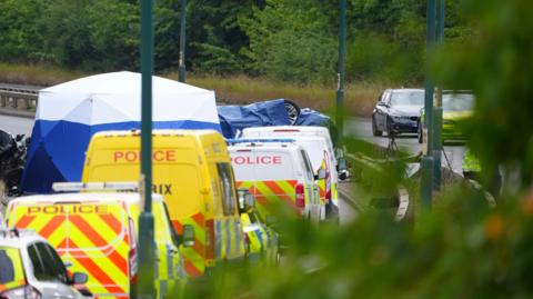 An overturned car is under a blue tarpaulin and surrounded by eight police vehicles on a road. There are bushes in the background and street lights along the central reservation which curves round to the left as it heads away from the camera.