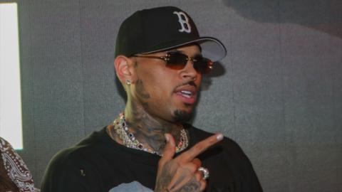 Chris Brown is wearing a black sweatshirt with a design printed on it in white. He is wearing a black baseball cap and shades.