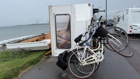 A caravan is blown in its side on a beachside road. It has two bikes attached to the back