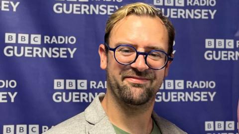 Gene Fallaize. he has short brown and blonde hair and short brown stubble. He is wearing large circular blue rimmed glasses. He is wearing a grey suit jacket and a green t shirt. He is stood in front of a purple BBC Guernsey banner.