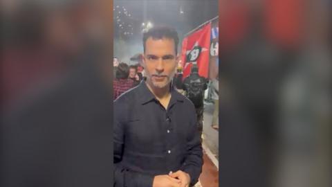 Man with black shirt standing in front of red flag with Lula's face