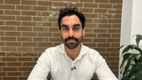 A young man with dark curly hair wearing a white shirt sits in an office with a plant and a brick effect wall behind him.