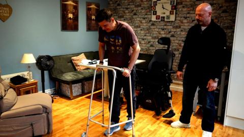 Keith Dungait using a walking frame. He has short dark hair and is wearing a brown t-shirt and dark trousers. There is a wheelchair behind him. To his left is Alan Shearer, wearing a dark fleece, dark trousers and white trainers. The pair are in a living room setting, with a sofa and armchair and and exposed brick wall behind them.