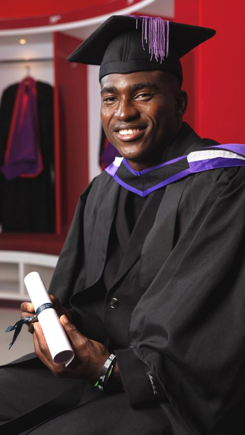 Taiwo Awoniyi wears a black robe with purple and white trim on the shoulders as well as a black mortar board with purple tassel dropping forward over the front. He smiles into the camera as he holds a white scroll tied up with a black ribbon while sitting in the red and white dressing room at Arsenal's Emirates Stadium