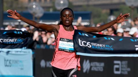 Peres Jepchirchir in a pink top with arms outstretched, breaking a ribbon as she crosses the finish line at the Sydney marathon