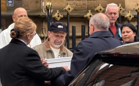 A man with a grey beard and wearing a black baseball cap and light brown jacket and cream sweatshirt appears to be crying as he holds a small white casket. Other mourners and a priest in a white cassock look on. Behind them are the black iron railings capped with gold coloured crosses and the stone wall of the cathedral.