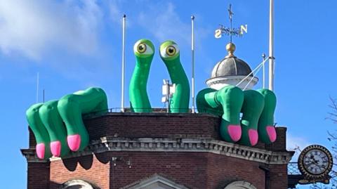 A green inflatable monster with eyes on stalks and claws hanging over the balustrades sits on top of Maidstone Town Hall
