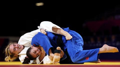 Emma Reid wearing white and Maria Swan in blue are judo fighting on the floor. Emma has Maria in an arm lock on her way to winning Commonwealth judo gold.