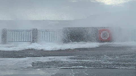 A picture of waves crashing onto Douglas Promenade on the Isle of Man. Standing water can be seen on the road with drops of water falling onto the surface through the railings of the wall.