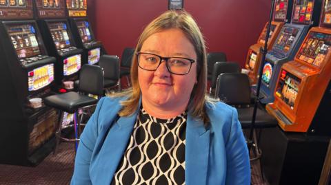 Sarah Calakovic sits in from of a row of slot machines. She is wearing glasses and a blazer.