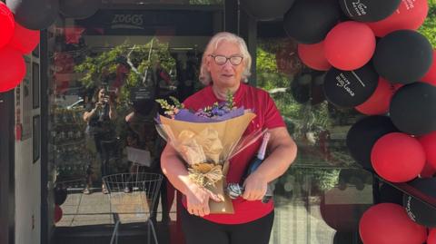 Diane is wearing a red top and is wearing lasses and holding a bunch of flowers. She is stood in the doorway to a leisure centre which has red and black balloons in an arch going over it.