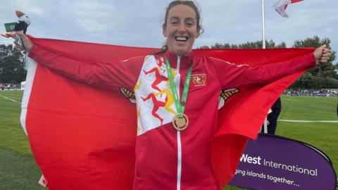 Rachel Franklin, wearing a red Isle of Man flagged jacket, with a gold meal, holding a Manx flag behind her.