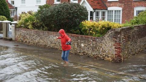 A woman in a red coat and jeans walks barefoot through a flooded road. The water comes up to her shins. Behind her is a flint wall and some pretty cottages with rose bushes and shrubs outside.