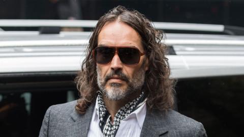 Russell Brand arriving at court on Monday 26 October - he is wearing a grey suit jacket, white shirt and black and white cravat. He is also wearing sunglasses.