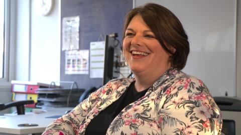 Charlotte Riley has shoulder-length dark brown hair and is wearing a white, pink and black floral jacket over a black top. She is sitting in front of a desk in what looks like an office and she is smiling widely to someone off camera.
