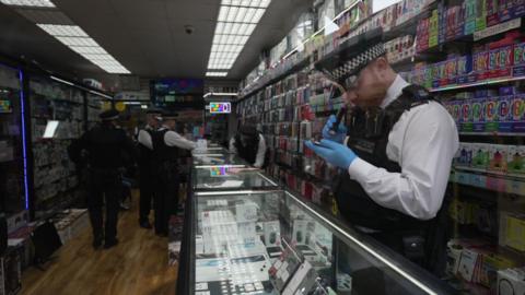 Metropolitan Police officers examine mobile phones and boxed electronic devices inside a Brixton phone shop during a raid targeting suspected stolen goods. Reflections in the shop window show officers checking stock displayed behind the counter.
