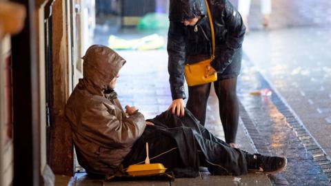 A passer by touches the knee of a homeless person sitting on the floor on a street as they check in with them, both hooded on a rainy day in Cardiff in 2018.