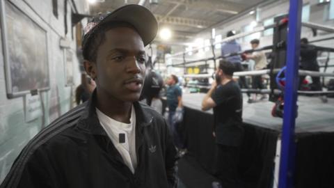 A young man wearing a bejewelled cap stands in front of a boxing ring at a youth club