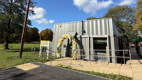 Grey metal building, a youth club, with a balloon arch and a sign outside - on a sunny autumn day in Wheatley near Doncaster. The sky is blue and there is an autumn tree on the right of the yellow pavement around the building.