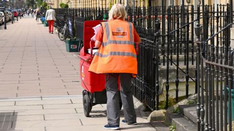 A postie delivers letters on a street, walking next to some railings.