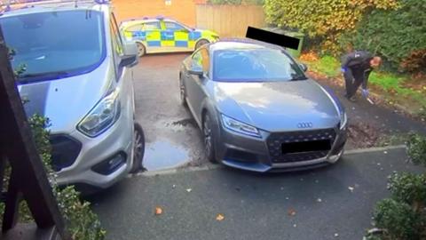 A police worker searches outside a property with a car and a van parked outside. A police car is in the background.