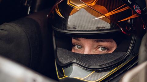 Swedish Top Fuel drag racing driver Susanne Callin wearing a black and orange racing helmet with just her eyes exposed through the visor, looking directly at the camera, taken at Mantorp in Sweden in 2025.