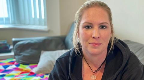 Jo, sitting in a room, by a sofa. She is looking at the camera and is wearing a black top, necklace and has a patterned blanket on the sofa. She has long blonde hair tied back.