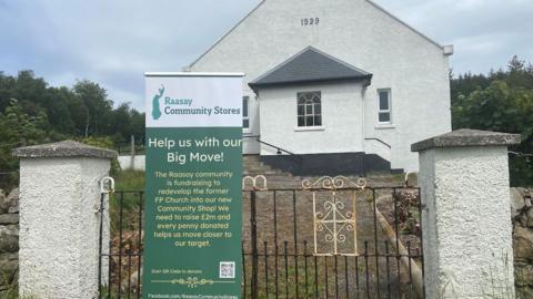 The church is a single-storey building with white harled walls. In the foreground is a metal gate between two white pillars and a white and green sign explaining the Raasay Community Stores project.