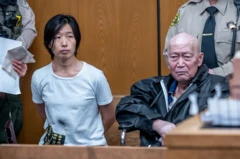 Silvia Zhang, vestida con una camiseta blanca y con las manos a la espalda, está sentada en una sala del tribunal junto a Guojun Xuan, un hombre calvo de mediana edad, con dos agentes de pie detrás de ambos.