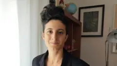 Sofía tiene el pelo corto y oscuro, peinado con un tupé. Lleva un blazer negro y está de pie en una habitación con una lámpara y una estantería al fondo.