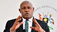 Minisitiri w'ubuzima wa Uganda Chris Baryomunsi arimo kuvugira mu ndangururamajwi. Akoze ikimenyetso n'ibiganza. Yambaye ikositimu y'umukara, ishati y'umweru na karuvati irimo amabara y'ubururu na khaki.