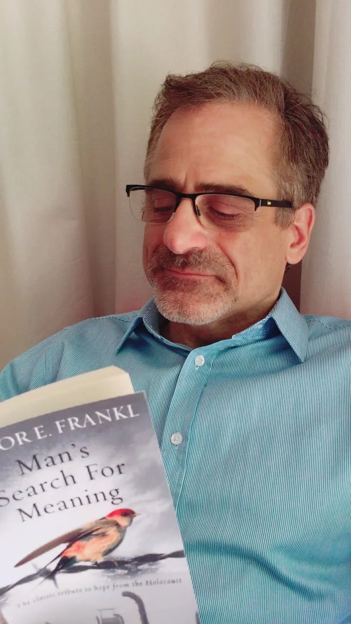 A middle-aged man with glasses and gray hair, wearing a light blue button-up shirt, reading a book titled 'Man's Search For Meaning' by Viktor E. Frankl, sitting against a curtain.