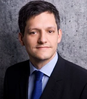 Headshot of a man in a suit and tie against a textured gray background.