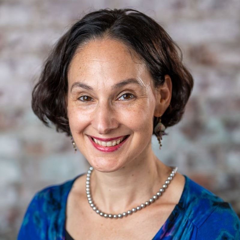 Smiling woman with short dark hair, wearing a blue dress, pearl necklace, and earrings, against a brick wall background.