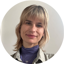 A woman with shoulder-length blonde hair, light makeup, and a slight smile, wearing a purple top with a beige jacket, standing against a plain light-colored background.