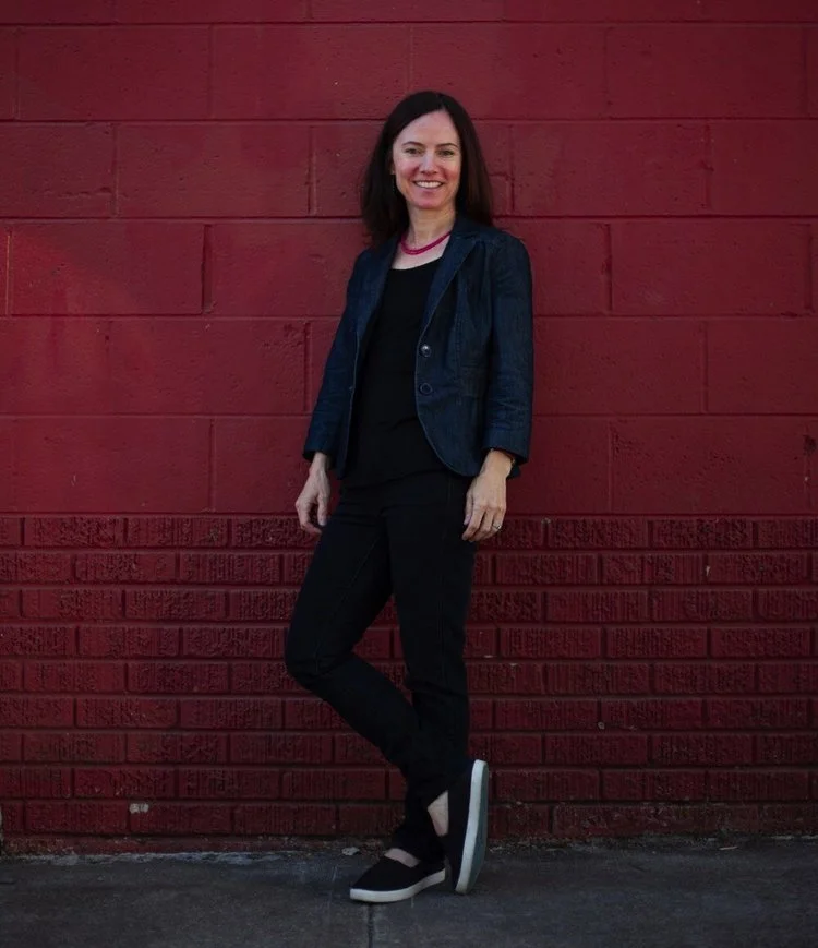 A woman standing in front of a red brick wall, smiling, wearing a dark blue blazer, black top, black pants, and black slip-on shoes.