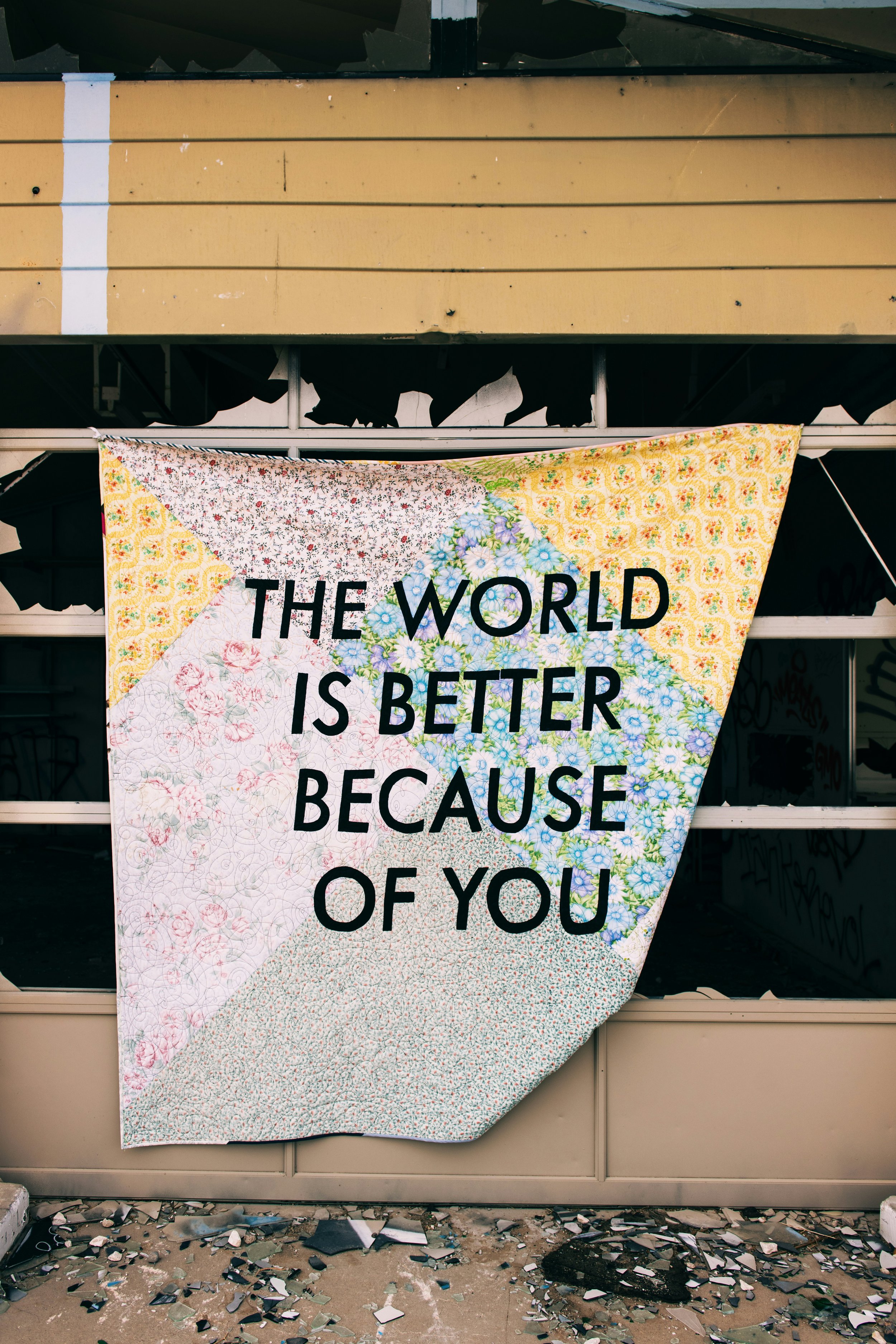 A quilt with floral patterns and geometric shapes hanging on a wall, with the words 'THE WORLD IS BETTER BECAUSE OF YOU' written on it.