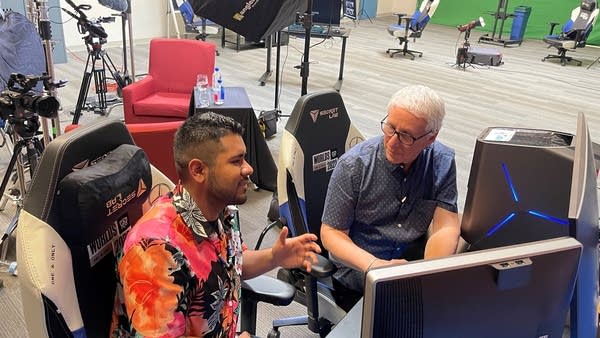 Rogelio Lara of Gameheads sits and talks with "Marketplace Morning Report" host David Brancaccio. They're in the video game development studio, in front of computers, with green screens and video equipment in the background.