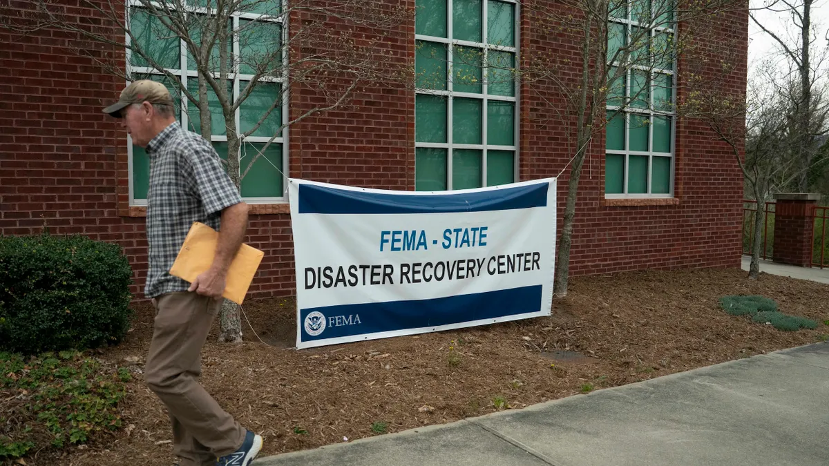 A person walks past a brick building and a banner hung between two trees that says "FEMA - STATE DISASTER RECOVERY CENTER."