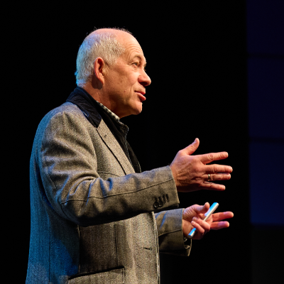 A person in a gray blazer speaks on stage, holding a pen. The background is dark.