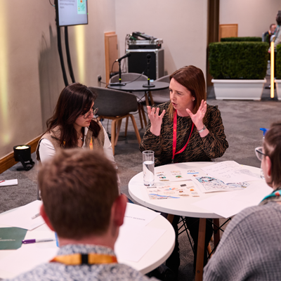 A group of people sit around a table in a conference room, discussing. Papers and a glass of water are on the table.