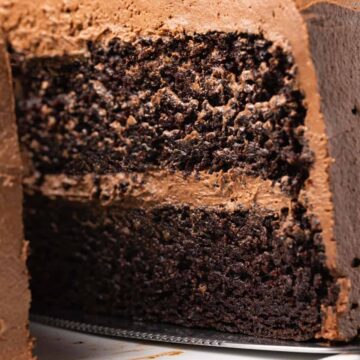 Vegan chocolate cake on a white cake stand with one slice cut and ready to serve.