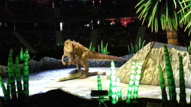 A realistic-looking Tyrannosaurus rex is propelled by a low, flat, grey car