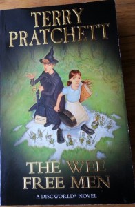 a book, The Wee Free Men. On the cover stands a young girl holding a big book and a frying pan. Beside her is a witch dressed in black holding a contraption of string, twigs, and stones. On the brim of her pointy hat sits a yellow toad. At their feet are several tiny blue men armed with swords. All except the witch are in an attitude of alarm and defence. The witch is watchful.
