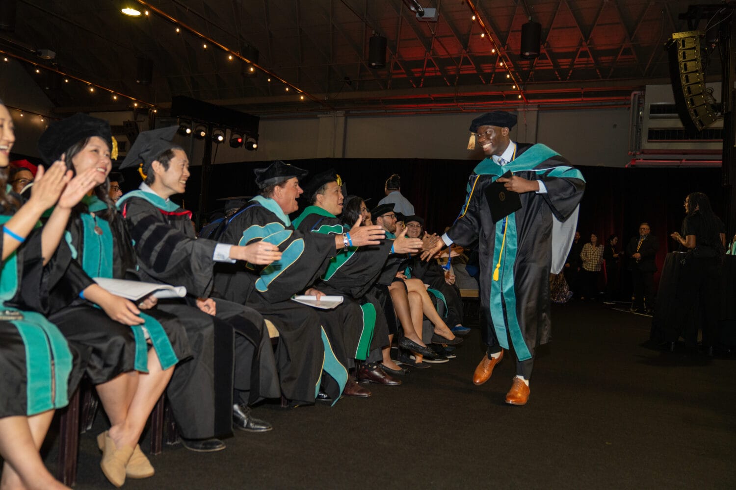 A graduate in cap and gown high-fives seated faculty members during a graduation ceremony inside a large indoor venue.